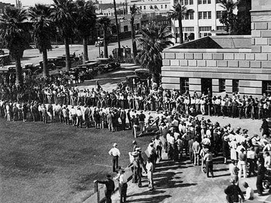 A black and white photo of people standing in front of buildings.